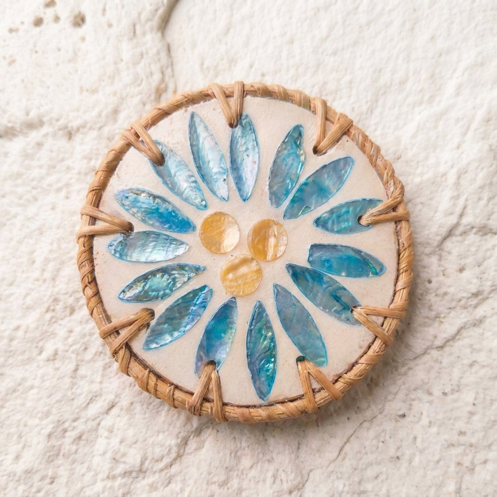 A round woven basket or wicker tray with a floral mosaic design featuring blue iridescent shell pieces and three small amber-yellow beads on a light beige background, set against a textured off-white stone surface by Bamboo Switch