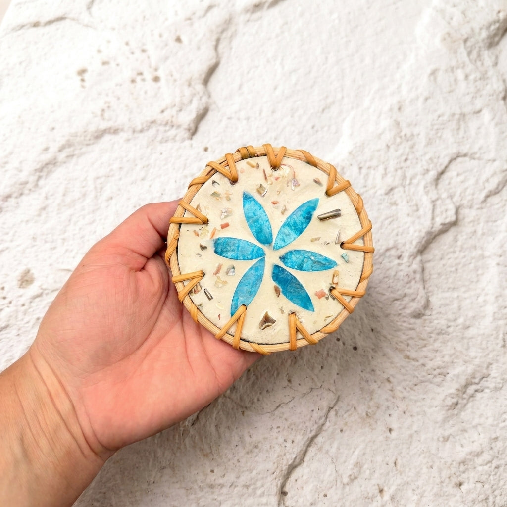A hand holding a round, handmade wooden bead craft piece featuring a blue six-petal flower design and metallic gold flecks on a textured cream background, set against a white stone wall by Bamboo Switch