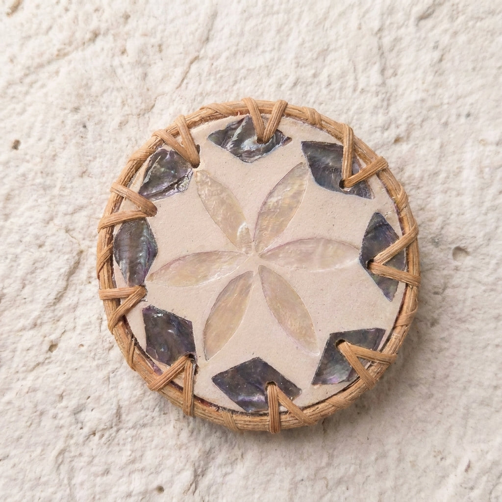 A round woven basket or tray with a natural tan wicker base, featuring a central beige floral pattern and alternating dark gray and black shell inlays around the edge, set against a textured off-white background by Bamboo Switch