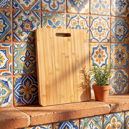 A rectangular bamboo cutting board with a handle cutout, leaning against a wall of colorful patterned ceramic tiles next to a small potted plant.