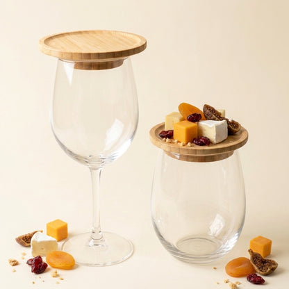 Two clear glass canisters with bamboo lids, one shaped like a wine glass and one rounded cube, styled with cheese, dried apricots, and figs on a warm cream background by Bamboo Switch