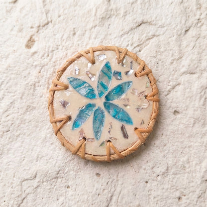 A round handmade ceramic tile coaster or mat featuring a blue and green floral starburst design on a beige background, surrounded by a braided tan border, set against a textured off-white wall by Bamboo Switch