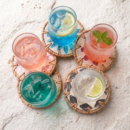 Four colorful cocktails in clear glasses with ice, arranged on woven round coasters on a light textured surface. The drinks are pink, blue, teal, and clear with lime or mint garnish by Bamboo Switch