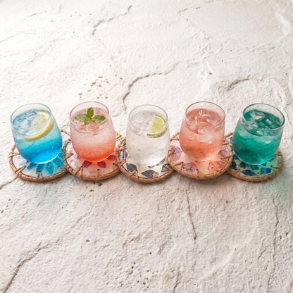 Five colorful cocktails on woven round coasters with floral mosaic detailing, arranged on a textured white surface by Bamboo Switch
