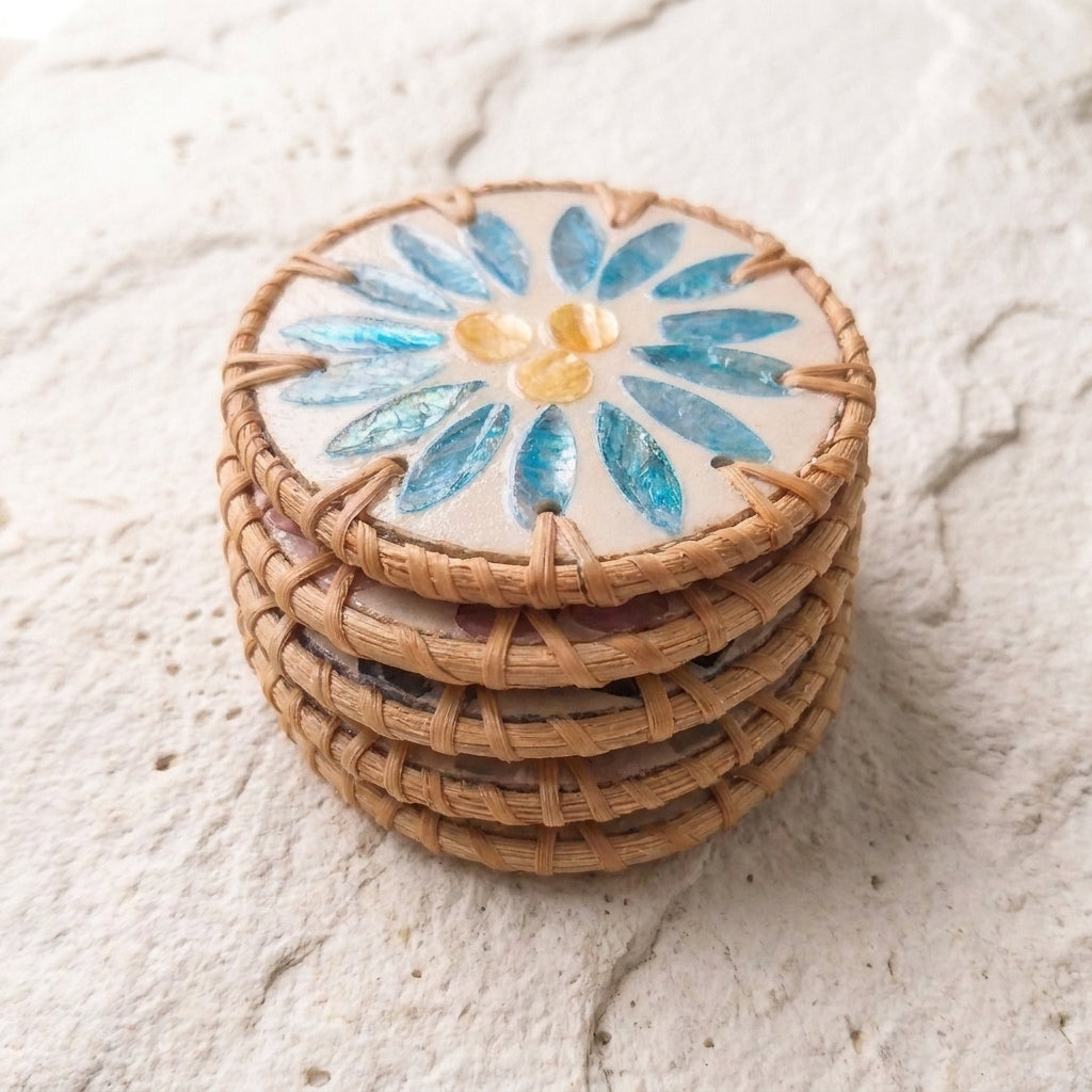 A stack of five round woven wicker coasters with a ceramic floral design on top, featuring blue and yellow petals on a cream background, set on a textured light beige stone surface by Bamboo Switch