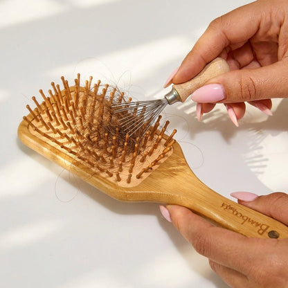 Bamboo hairbrush with bristle head held by a hand on a light background using a wire cleaning tool to remove hair