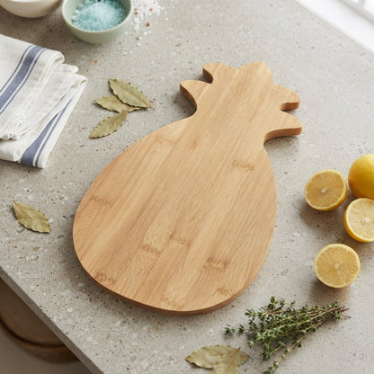 A bamboo cutting board shaped like a pineapple sits on a light speckled countertop, surrounded by lemons, bay leaves, herbs, and a folded cloth. No person or animal is present, and the scene is styled as a clean product setup rather than a real lived-in environment, so this fits Product/Packaging rather than Lifestyle.