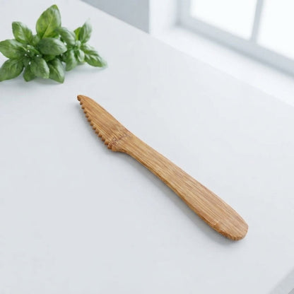 A light brown bamboo butter knife with serrated edges, resting diagonally on a white table next to a small green basil plant.