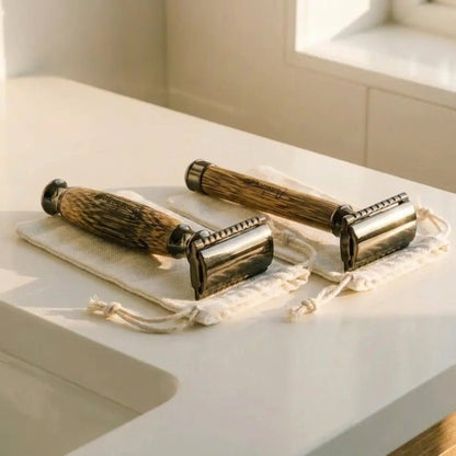 Two vintage-style metal safety razors with dark bamboo handles rest on small cream drawstring pouches on a white bathroom counter near a window by Bamboo Switch