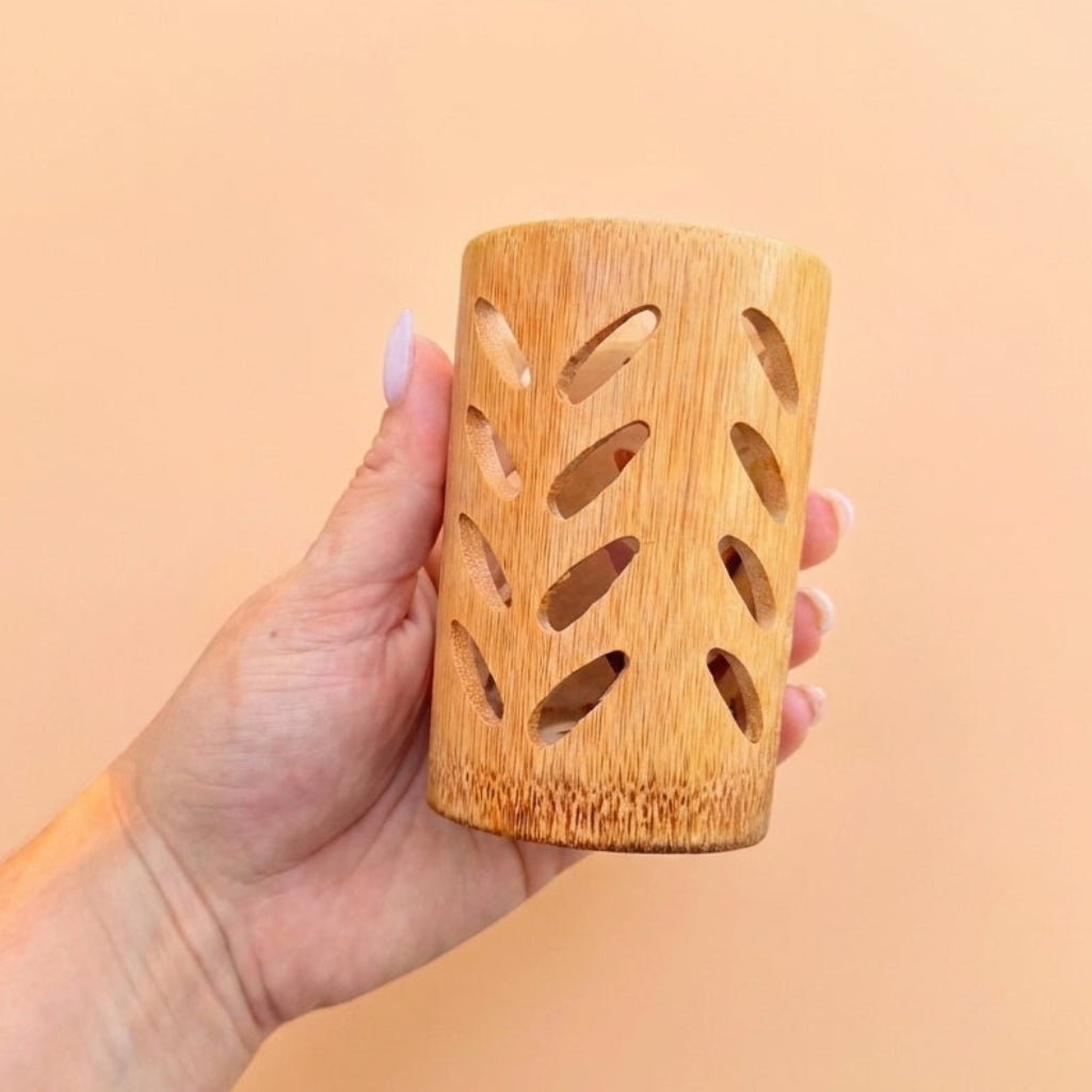A hand holding a small cylindrical bamboo holder with decorative leaf-shaped cutouts, set against a solid peach-colored background by Bamboo Switch