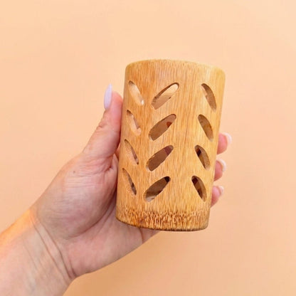 A hand holding a small cylindrical bamboo holder with decorative leaf-shaped cutouts, set against a solid peach-colored background by Bamboo Switch