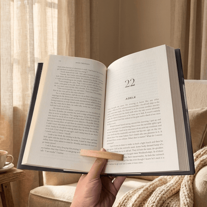 An open hardcover book held in a hand with a bamboo thumb book page opener on a cozy beige couch, with warm curtains and a cup of coffee in the background. The book pages are open to a chapter titled 'ADELE' and show the interior text by Bamboo Switch