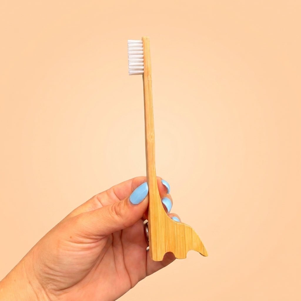 A hand with light blue nail polish holds a bamboo toothbrush with a dinosaur shaped handle against a solid peach background by Bamboo Switch