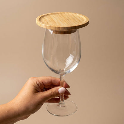 A hand holds a clear wine glass with a round bamboo lid resting on the rim, set against a solid beige background by Bamboo Switch