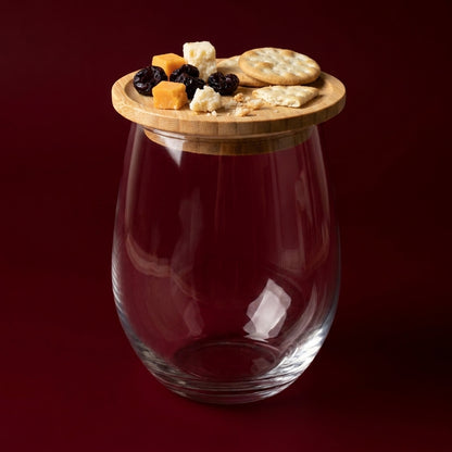 A clear glass stemless wine glass with a round light wood lid, shown filled with snacks including crackers, cheese cubes, and dark berries against a deep burgundy background by Bamboo Switch