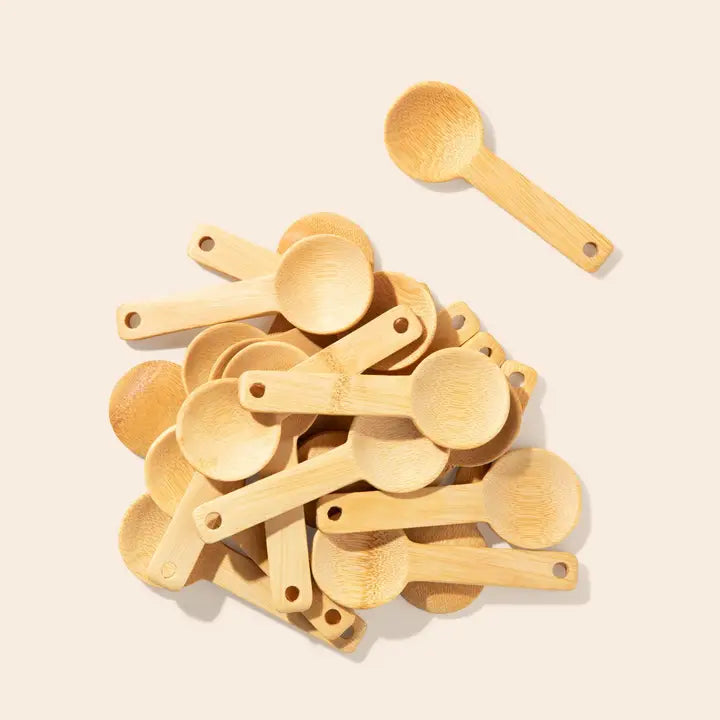 A pile of small bamboo measuring spoons with attached handles, shown on a plain light beige background. One spoon is placed above the pile, and the spoons are arranged in a clean, studio-style composition by Bamboo Switch