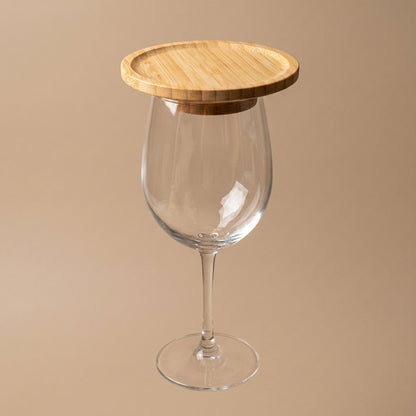 A clear wine glass with a round light wood lid resting on its rim, set against a solid beige background by Bamboo Switch