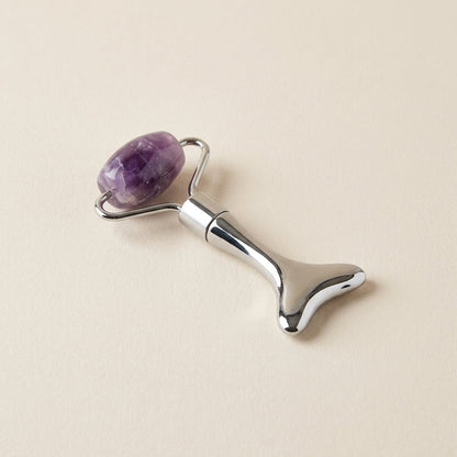 A silver metal facial roller tool with a polished purple amethyst stone roller head, shown on a light beige background by Bamboo Switch