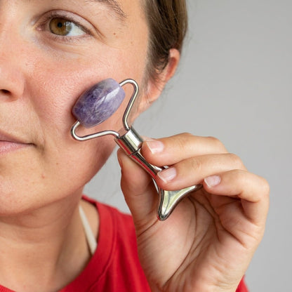 A close-up of a woman using a silver metal facial roller with a purple stone head on her cheek, wearing a red top against a neutral gray background by Bamboo Switch