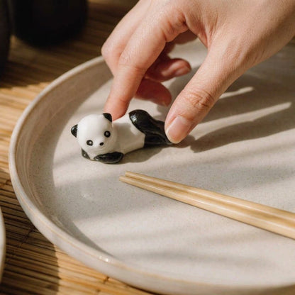 A small panda figurine resting on a beige ceramic plate, with a person's hand reaching toward it and bamboo chopsticks nearby by Bamboo Switch