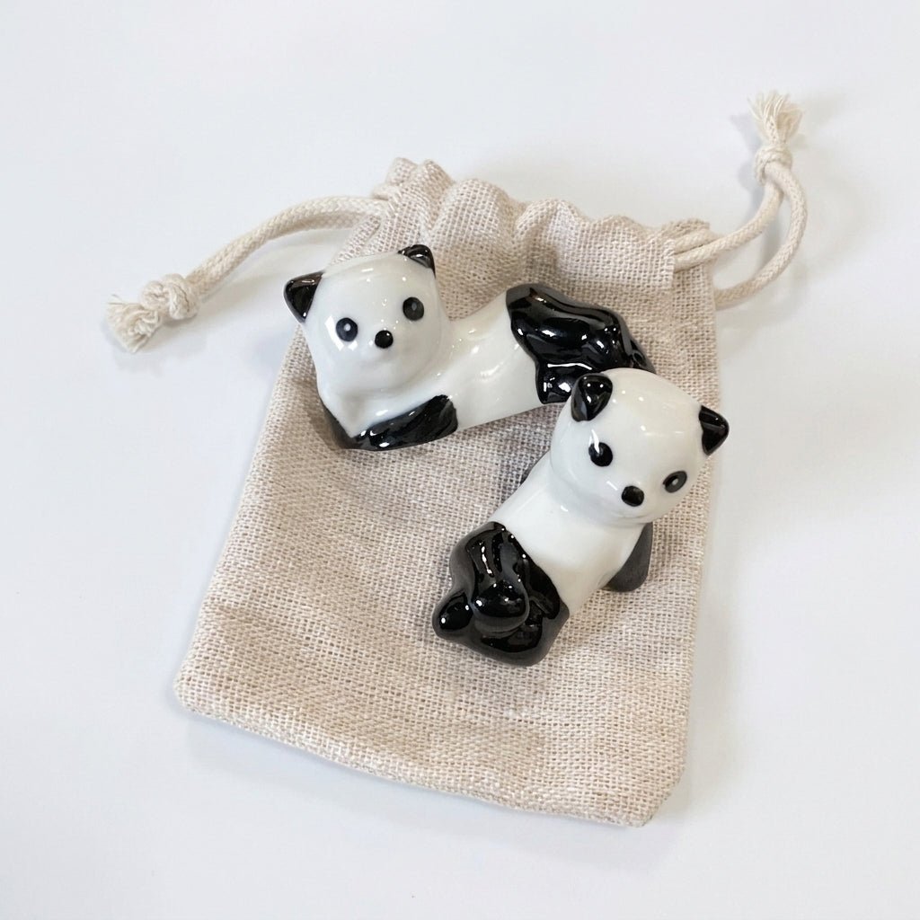 Two small panda figurines with glossy black and white coloring, displayed on top of a beige drawstring pouch against a plain white background by Bamboo Switch