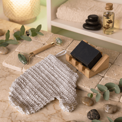 A spa-themed product collection featuring a textured off-white sisal glove, a bamboo-handled jade roller, a bar of charcoal soap on a bamboo stand, and green eucalyptus leaves on a beige stone surface by Bamboo Switch®